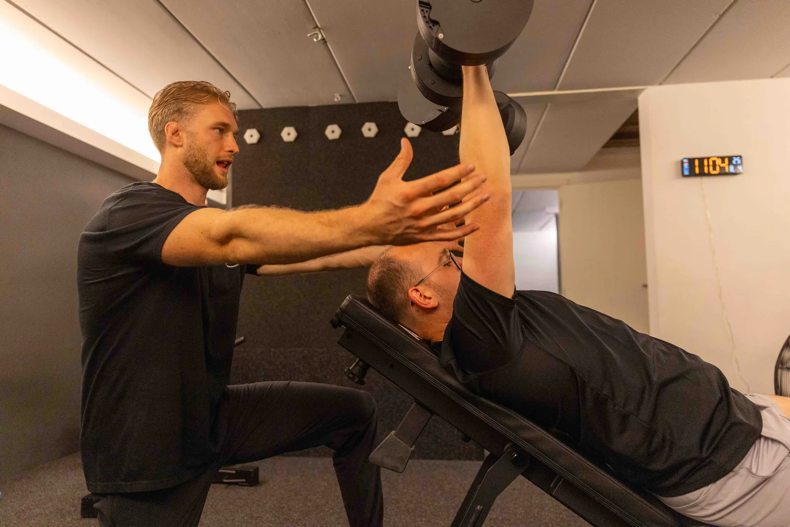 Robbe Berger begeleidt een klant bij een liggende dumbbell press op de bank