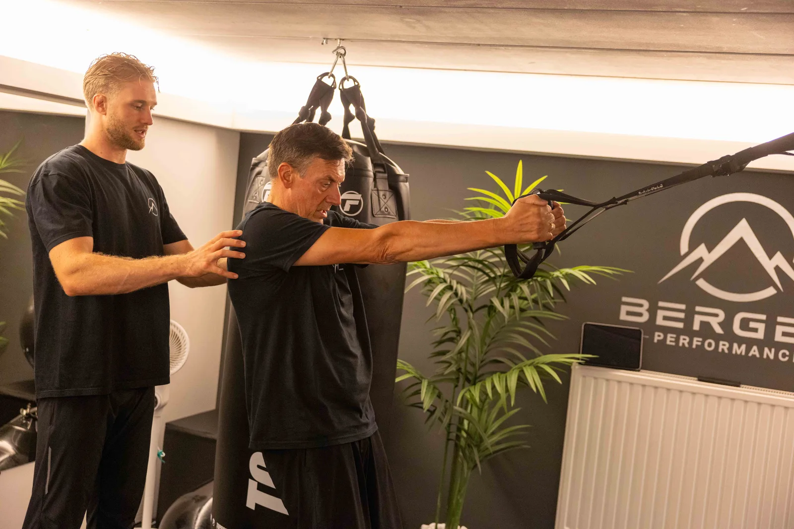 Robbe Berger coacht een klant aan de kabeltrainer, met bokszak en Berger Performance logo op de achtergrond
